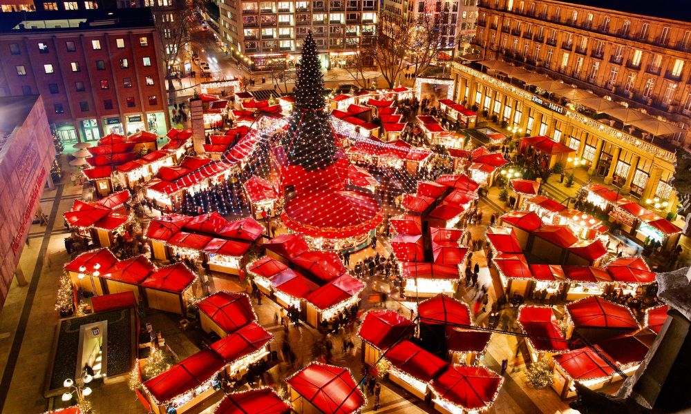 A festive display of German Christmas pastries and gingerbread at a stall in the Cologne Christmas market, with the Cologne Cathedral illuminated in the background.