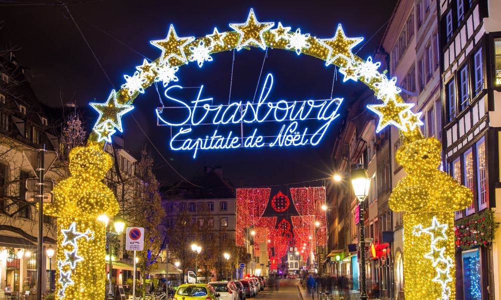 An assortment of French Christmas pastries and cookies at a stall in the Strasbourg Christmas market, with festive decorations and a large Christmas tree in the background.