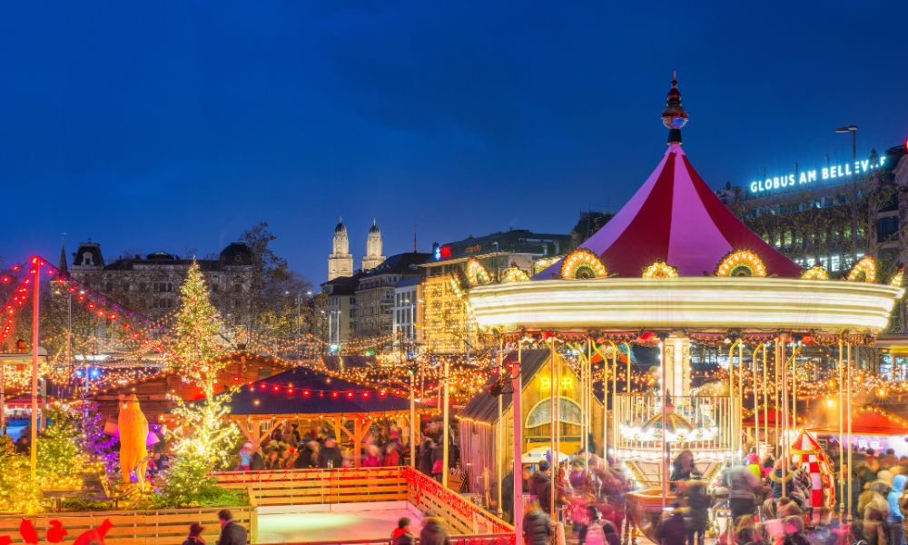 A display of traditional Swiss Christmas pastries and chocolates at a stall in the Zurich Christmas market, with festive lights and a Christmas tree in the background.