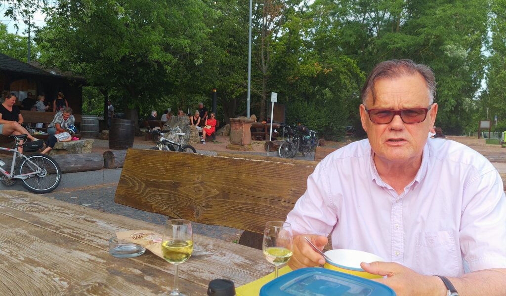 Hans enjoying a glass of wine at a Weintreff in Rheingau, with large wine barrels converted into wine bars and a view of the Rhine