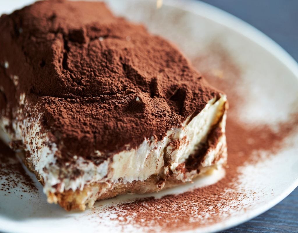 A perfectly layered slice of authentic Italian Tiramisu, topped with cocoa powder and dark chocolate shavings, served on a white plate